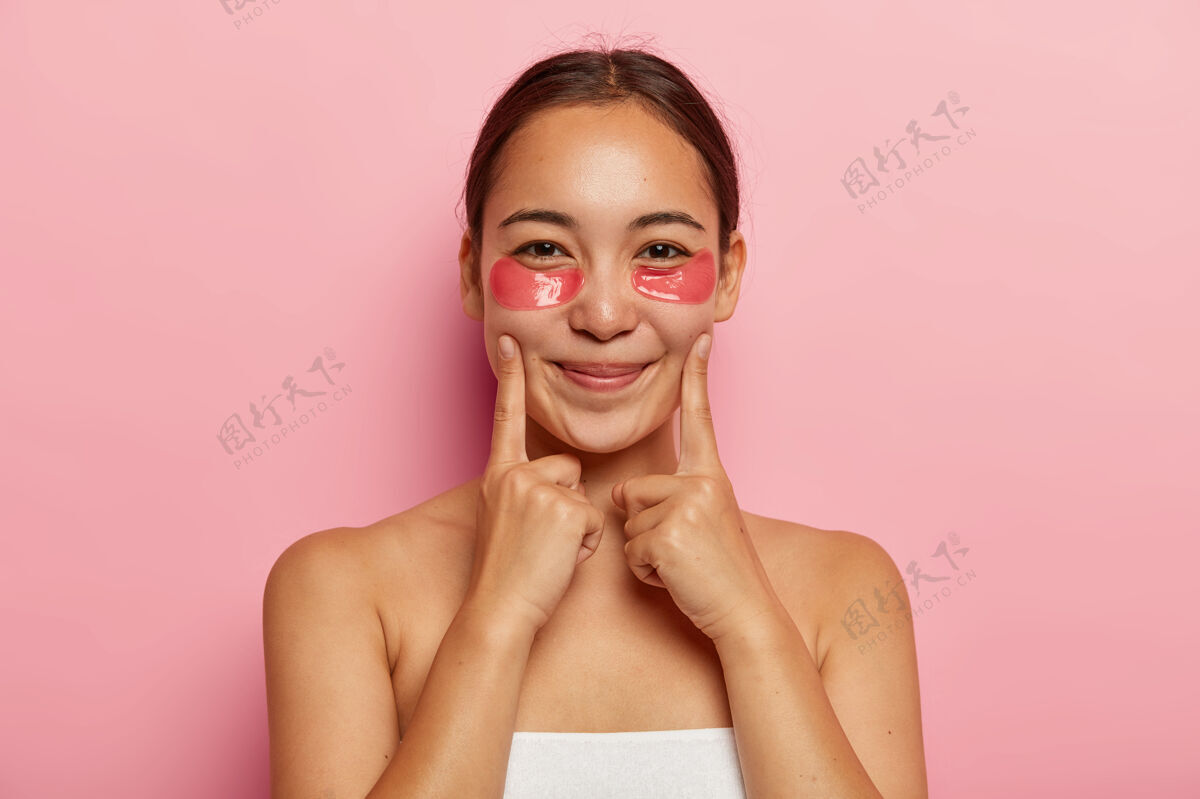 治疗美丽的韩国女性的特写肖像戴着眼袋下的化妆品 保持食指在脸颊上 微笑温柔 赤膊上阵 避免黑眼圈和皱纹在脸上模特脸颊姿势