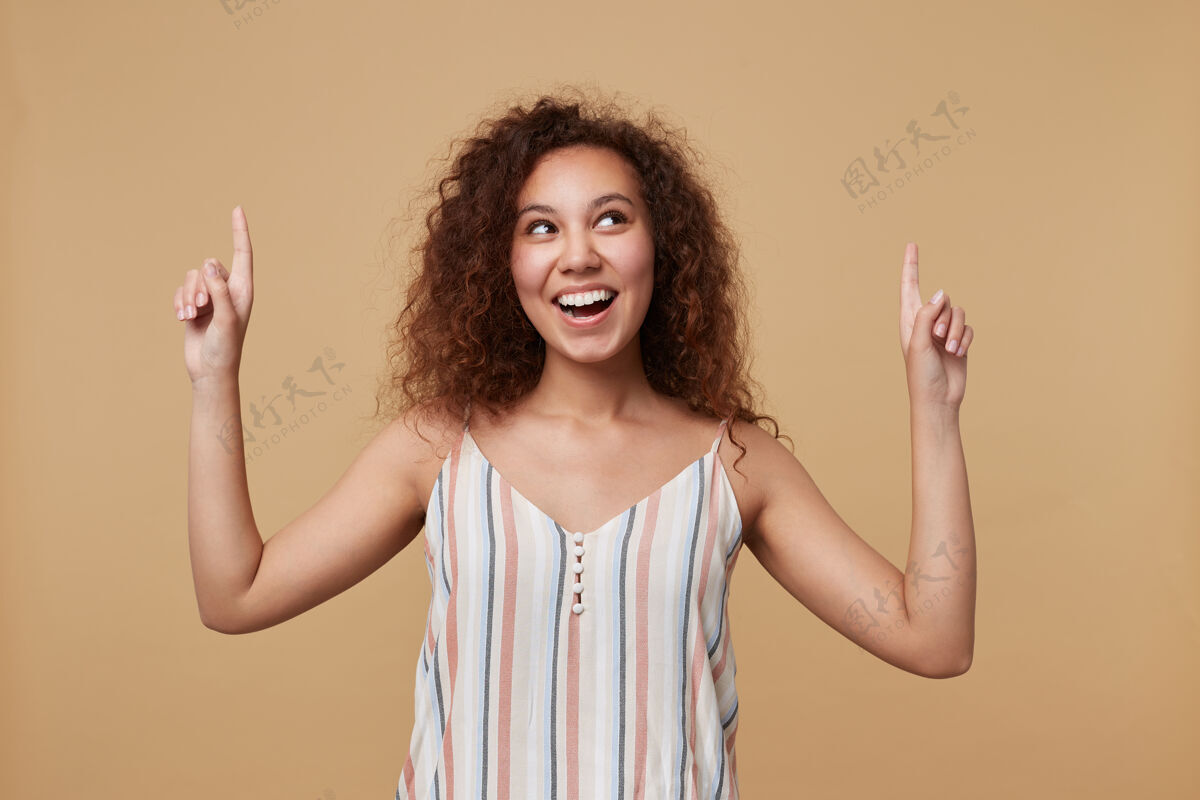 女士兴奋的年轻可爱的棕色头发卷曲的女人 穿着夏季条纹上衣 愉快地向上指 食指抬起 面带笑容 孤立地站在米色上正面表情女性