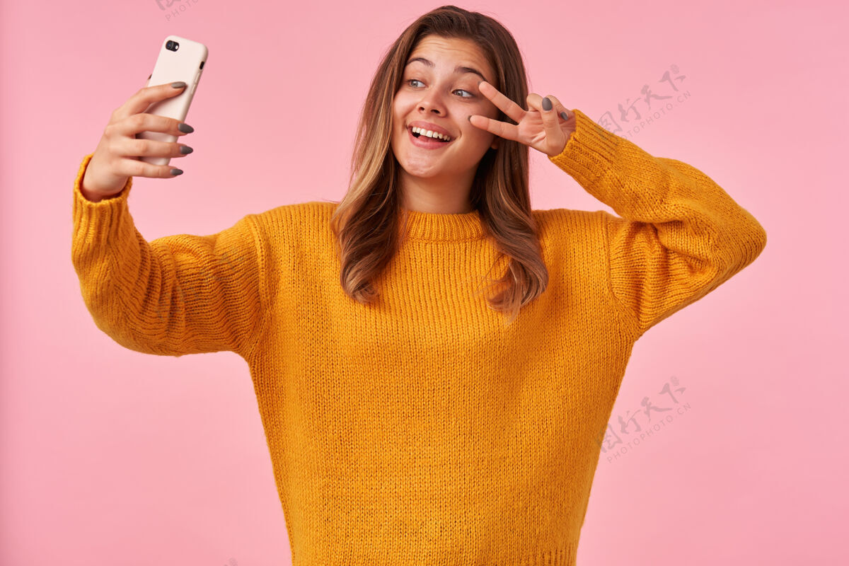 智能手机快乐的年轻漂亮的棕色头发的女性举起手 脸上挂着胜利的标志 在智能手机上为自己画像 穿着休闲服站在粉红色的地板上休闲手机舒适