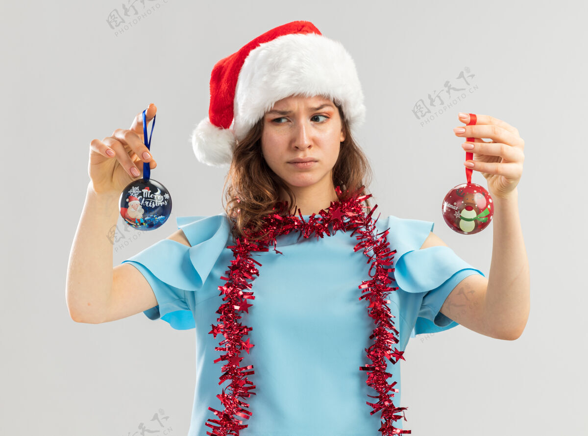 尝试一位身穿蓝色上衣 头戴圣诞帽 脖子上戴着金属箔的年轻女子 手里拿着圣诞球 看上去很困惑 试图做出选择圣诞老人金属片困惑