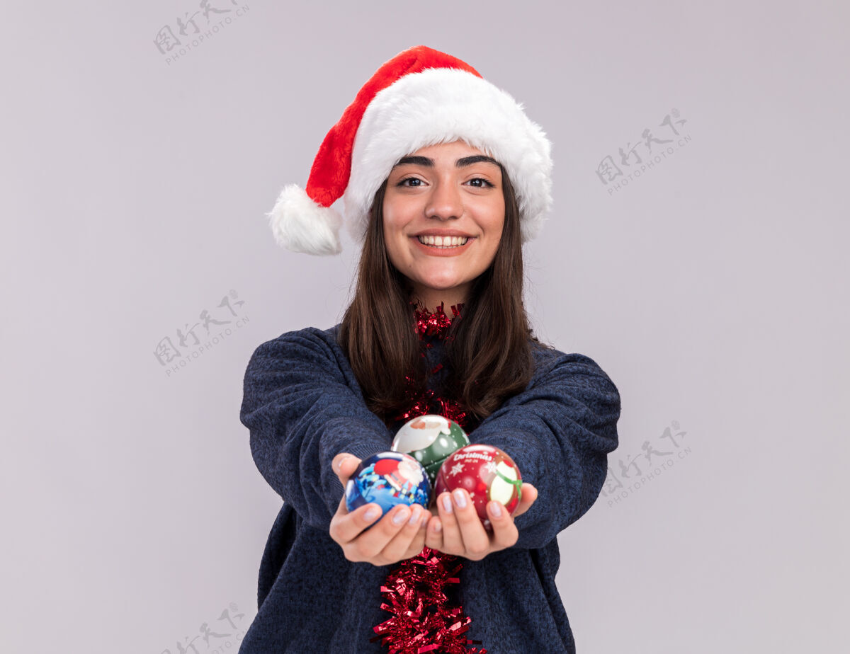 年轻微笑的年轻白人女孩戴着圣诞帽 脖子上戴着花环 手里拿着玻璃球饰品球帽子圣诞