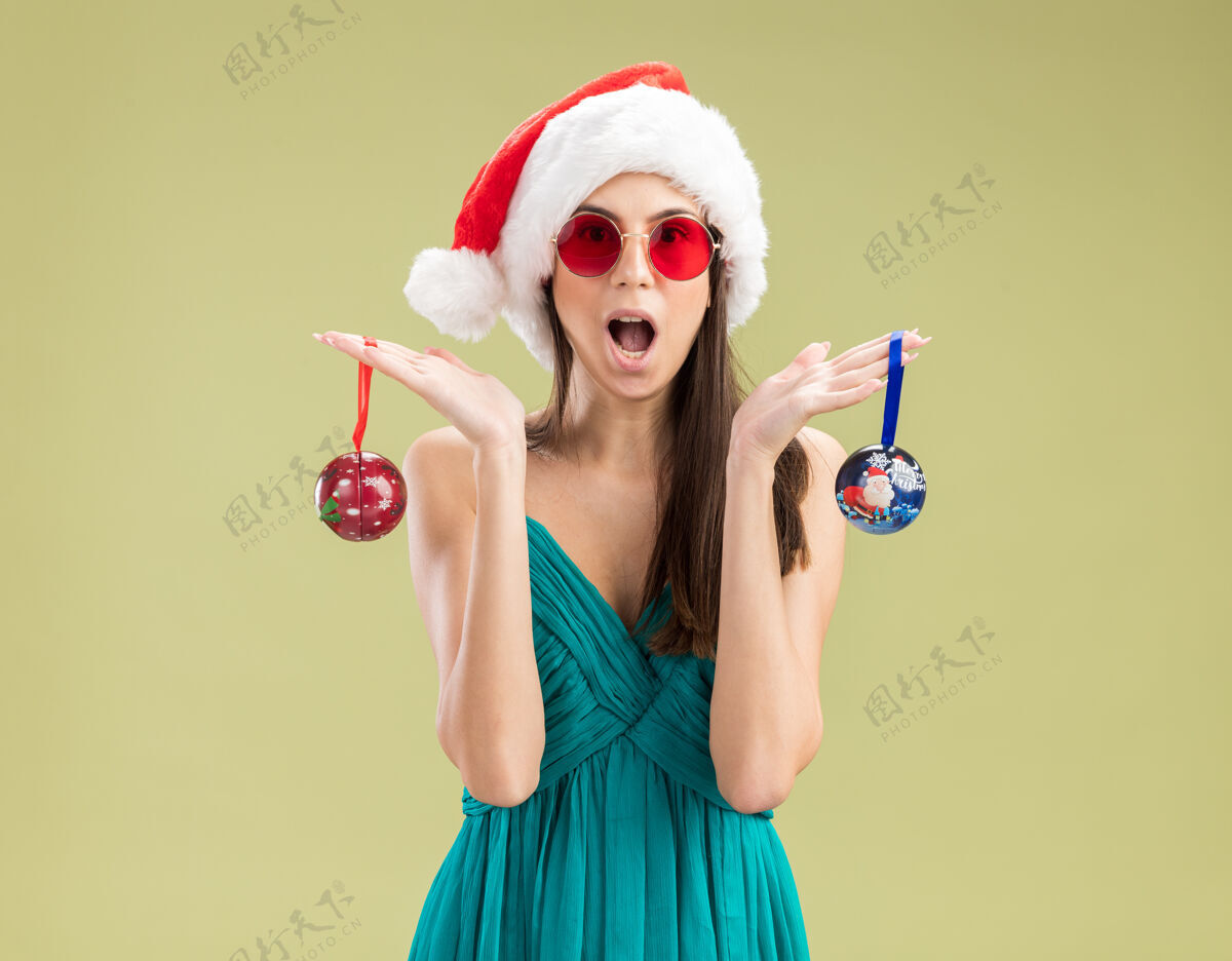 举行兴奋的年轻白人女孩戴着太阳眼镜 戴着圣诞帽 手里拿着玻璃球饰品新女孩装饰品