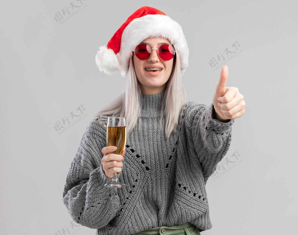 年轻年轻的金发女郎 穿着冬衣 戴着圣诞帽 手里拿着一杯香槟 看上去很开心 脸上露出积极的笑容 竖起大拇指圣诞老人帽子快乐