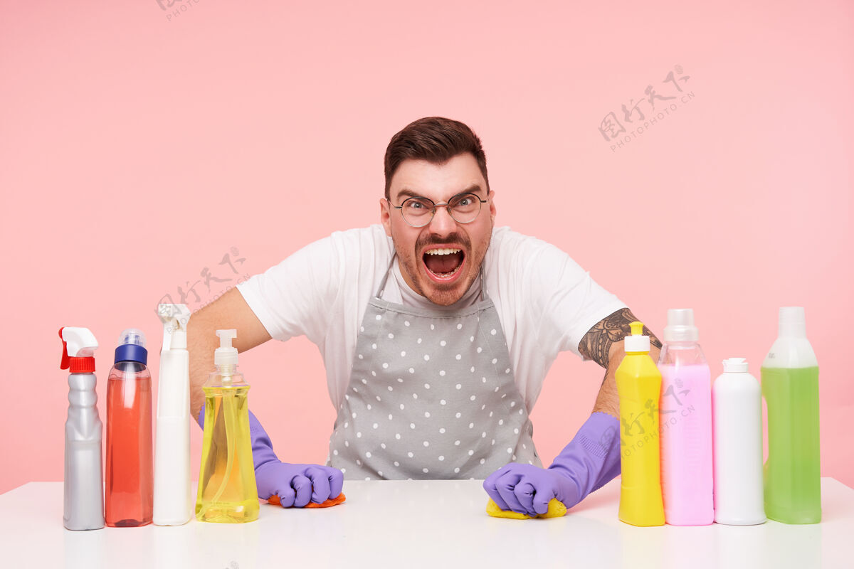 皱眉恼怒的年轻黑发戴眼镜的男人在打扫完房子后很累 在洗桌子的时候愤怒地尖叫 孤立在粉红色上清洁肖像男人