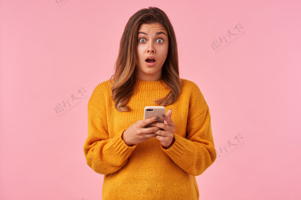 智能手机睁开眼睛的棕色头发的年轻女性 自然的妆容环绕着她的眼睛 当她在智能手机上读到意外的新闻后 惊讶地看着 孤立在粉红色上长款迷茫棕色