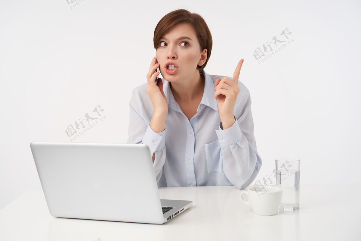 棕色室内画像 年轻的黑发女士留着时髦的短发 一边举起食指一边紧张地打电话 一边穿着蓝色衬衫一边摆着白色的姿势女性智能手机深色
