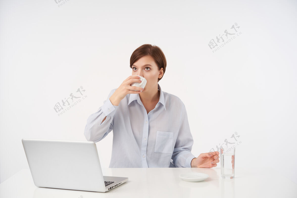 坐着室内画像年轻的棕色眼睛短发黑发女性与自然化妆喝咖啡 而在现代办公室与她的笔记本电脑 隔离在白色黑发陶瓷模特