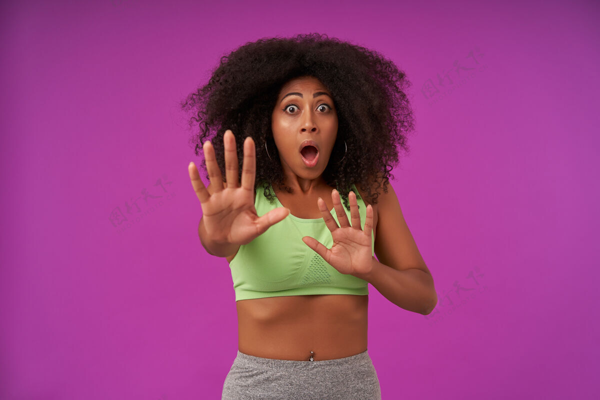 女性一幅受惊的年轻黑皮肤女人的画像 她卷发 穿着紫色的运动服 举手示意上衣黑色纽扣