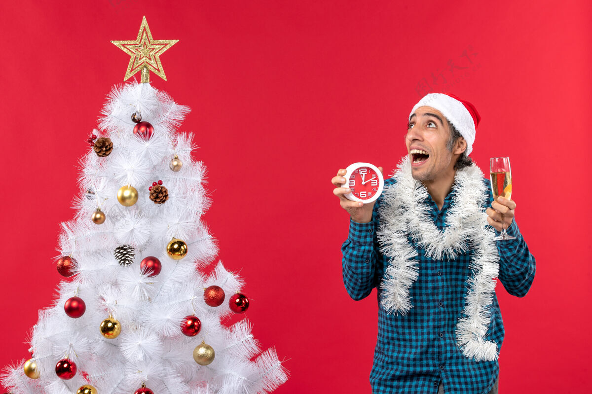 圣诞树一个戴着圣诞老人帽子 拿着一杯葡萄酒 站在圣诞树旁的闹钟的年轻人大吃一惊年轻人圣诞老人庆祝