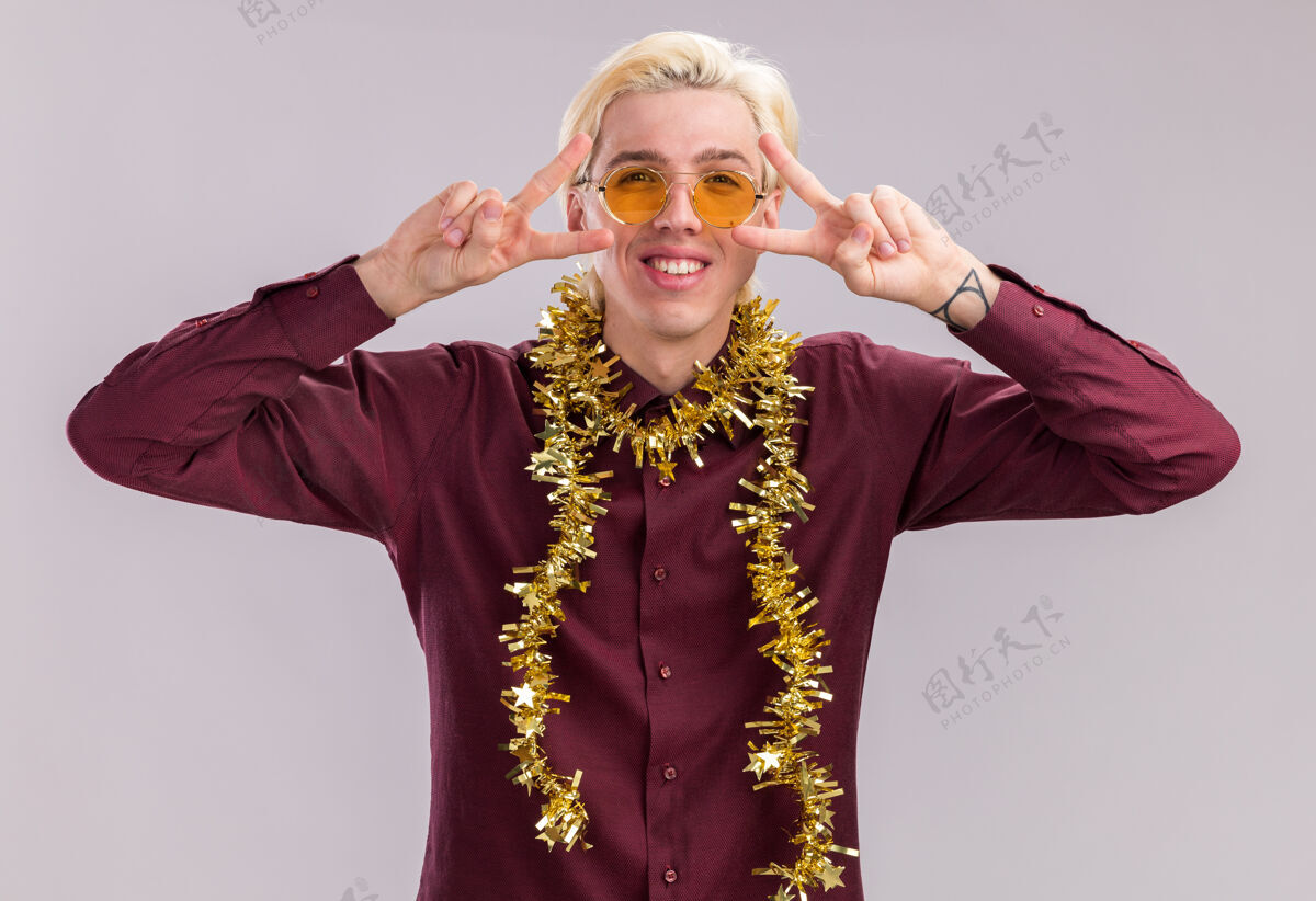 穿着微笑着的年轻金发男子戴着眼镜 脖子上戴着金属丝花环 在隔离在白墙上的眼睛附近显示着v符号花环近男人