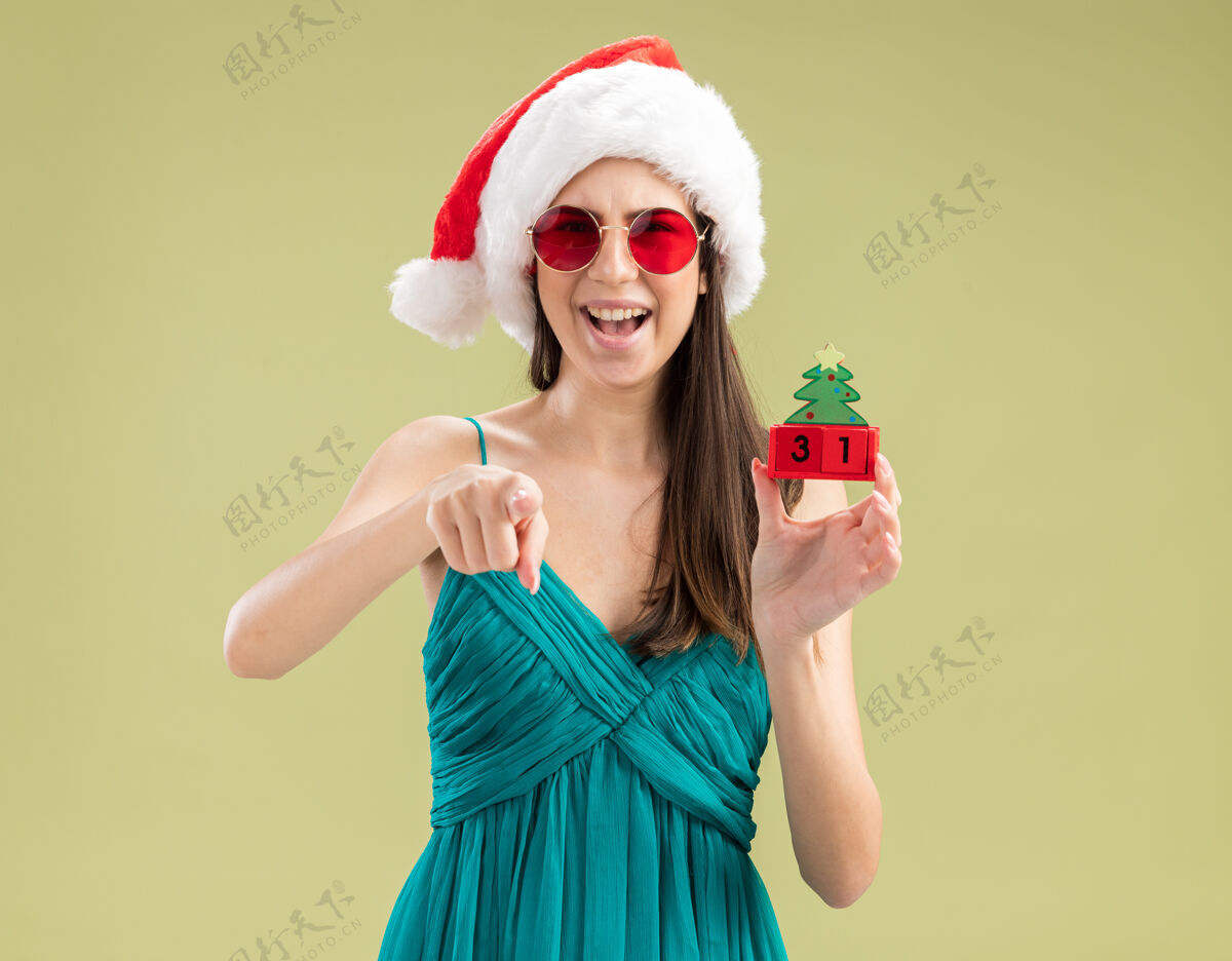 复制戴着太阳眼镜 戴着圣诞帽 拿着圣诞树装饰品和指头的快乐的年轻白人女孩圣诞快乐绿色新年