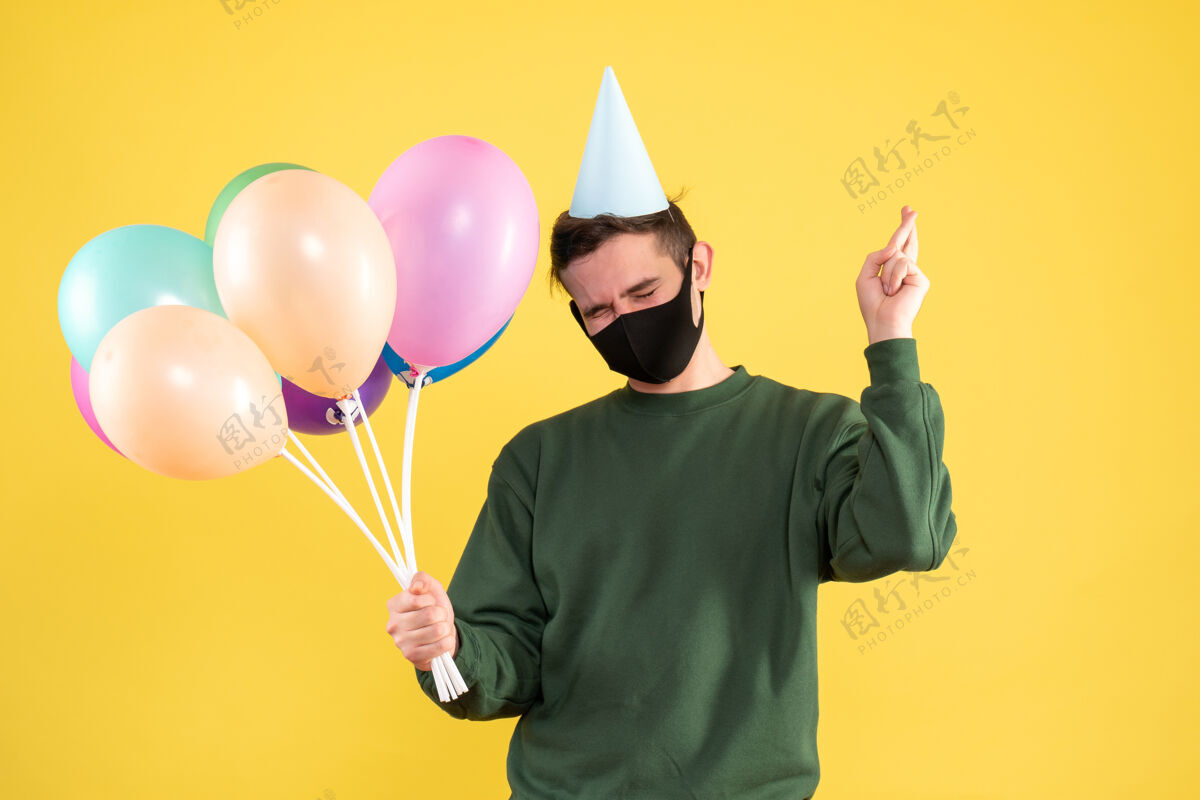 男人正面图：戴着派对帽的年轻人和五颜六色的气球 站在黄色的舞台上做着好运的手势五颜六色手制造