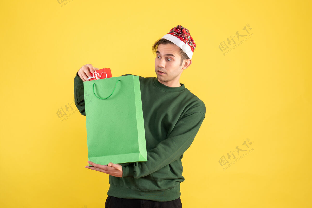 抱着正面图：戴着圣诞帽的年轻人拿着绿色的购物袋和礼物站在黄色的地板上好奇男人帽子