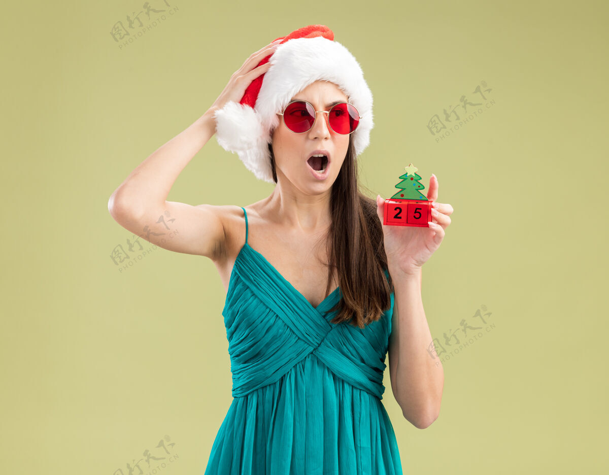 头一个戴着太阳眼镜 戴着圣诞帽的年轻白人女孩把手放在头上 手里拿着圣诞树饰物看着身边复制装饰品圣诞树