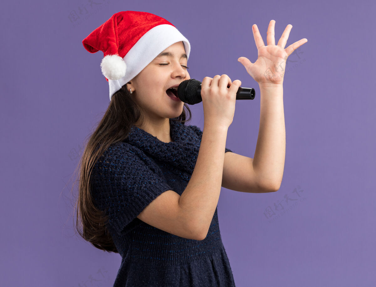 庆祝小女孩穿着针织连衣裙戴着圣诞帽拿着麦克风唱歌庆祝圣诞晚会快乐和积极新年紫色举行