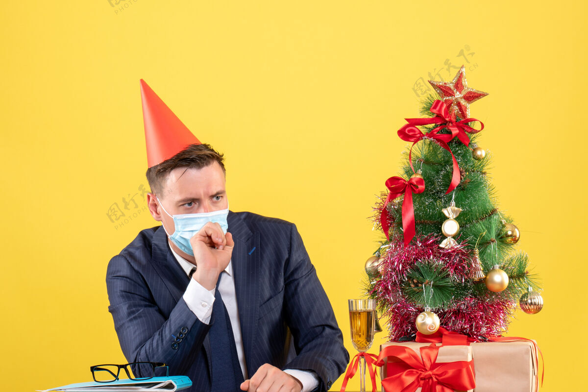 肖像带着医用面具的体贴男人坐在圣诞树旁的桌子前 黄色的礼物桌子公司黄色