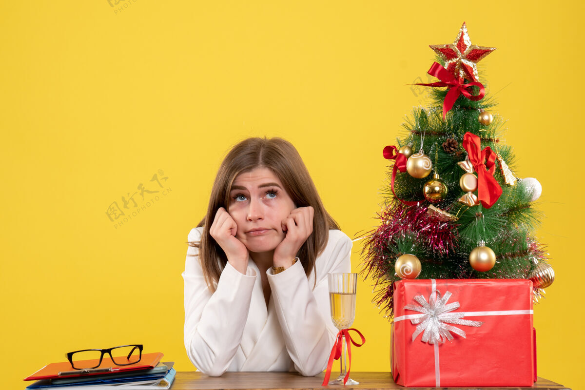 桌子正面图：女医生坐在黄色背景的桌子前 放着圣诞树和礼品盒前面快乐圣诞节
