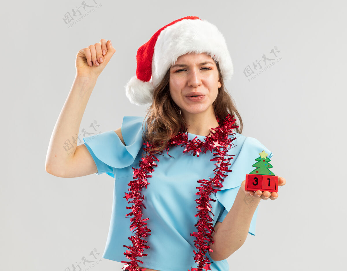 举行穿着蓝色上衣 戴着圣诞帽 脖子上戴着金属丝的年轻女子 手里拿着玩具立方体 手里握着快乐的新年约会的拳头 快乐而兴奋年轻帽子立方体
