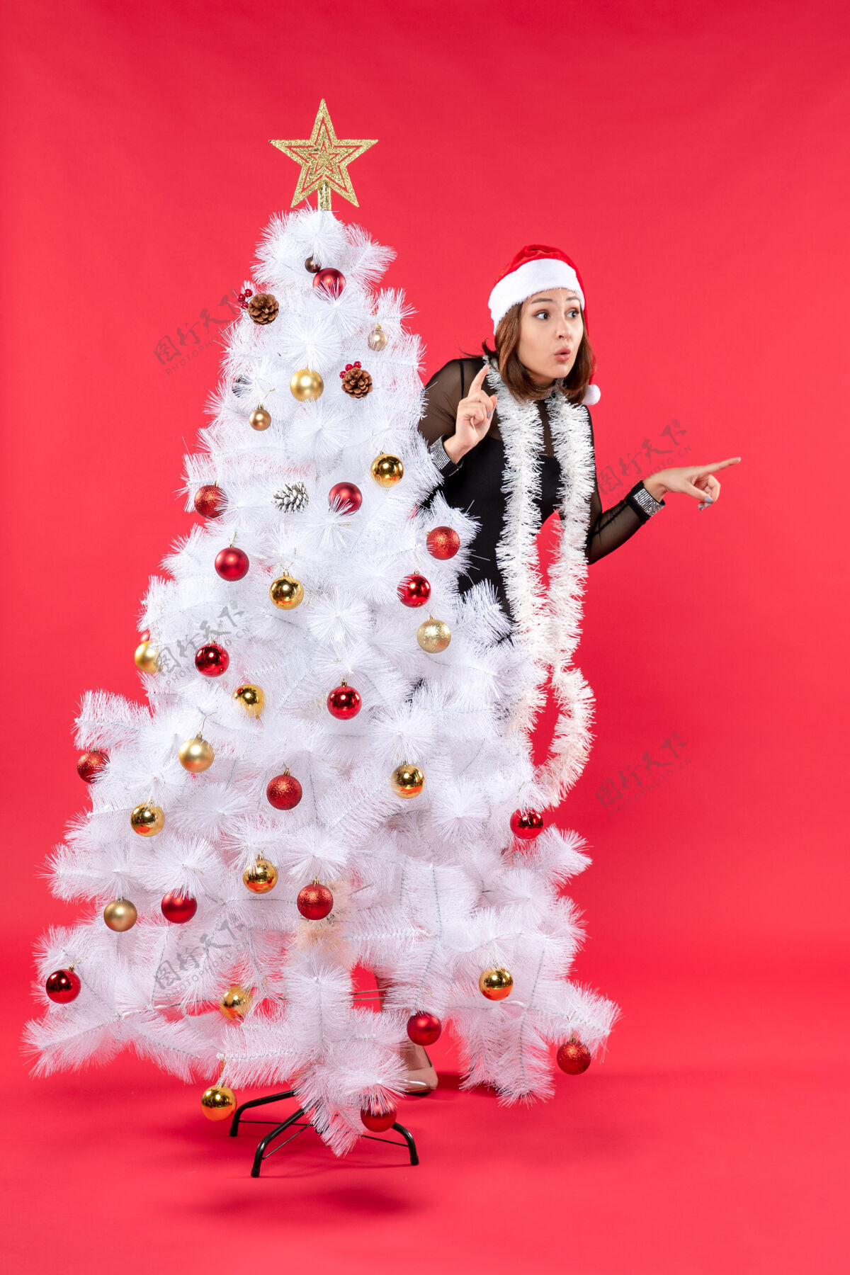 帽子圣诞心情与惊喜美丽的女孩在一个黑色礼服与圣诞老人的帽子隐藏在新年树股票照片庆祝衣服圣诞老人
