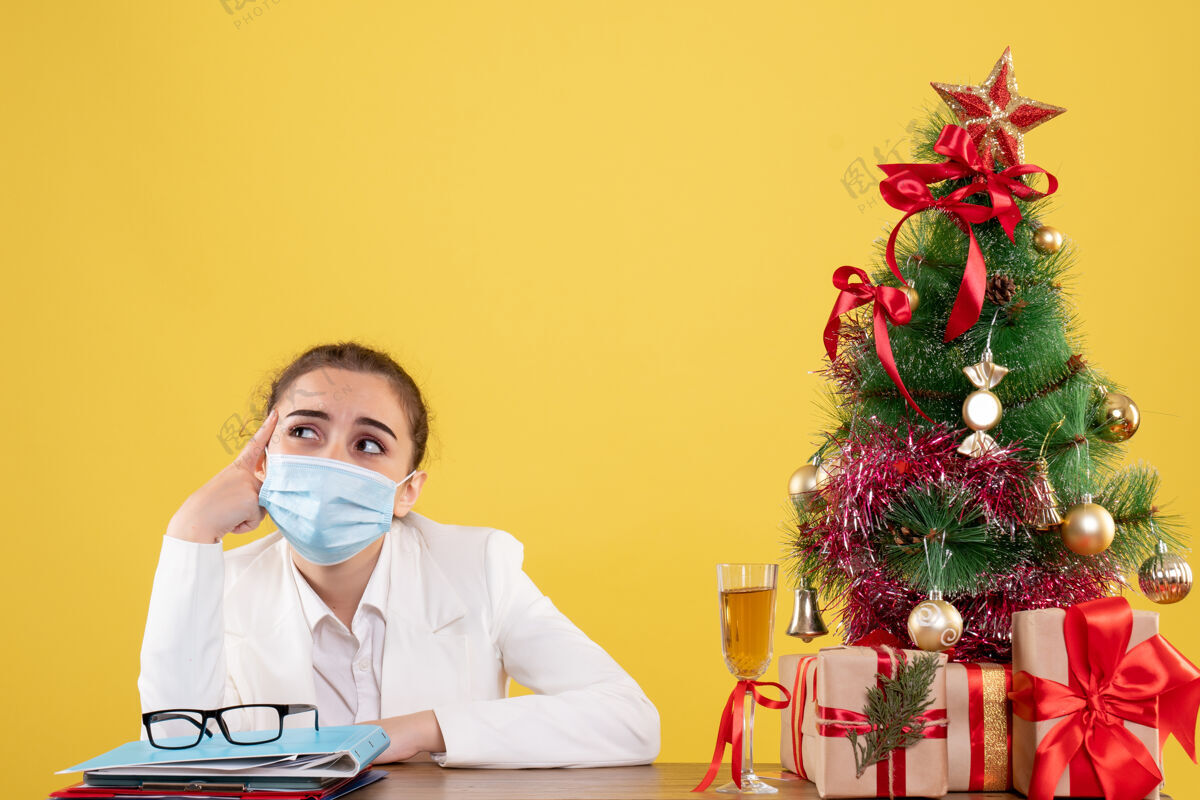 情感正面图女医生戴着无菌口罩坐在黄色背景上 带着圣诞树和礼品盒实验室医疗背景
