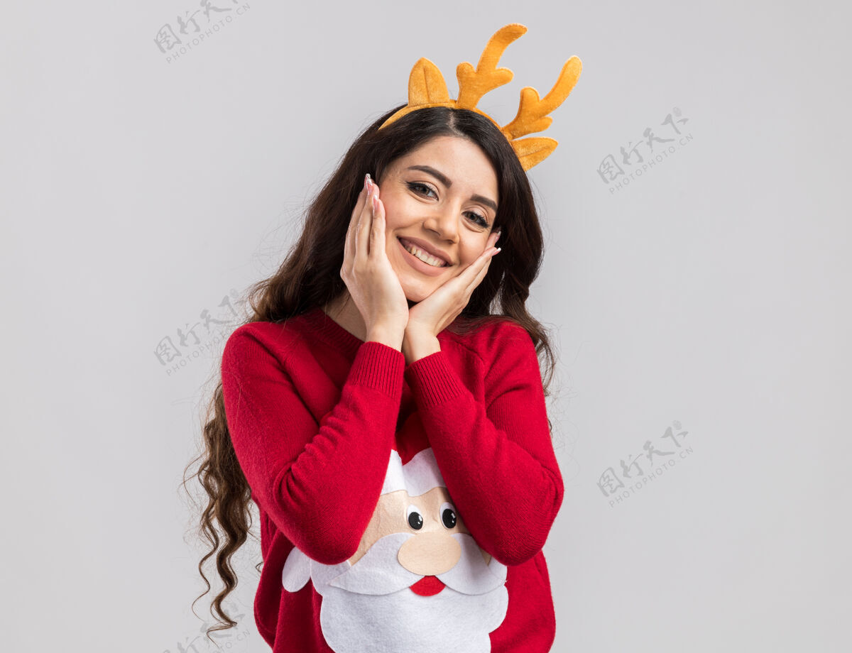 毛衣微笑着的年轻漂亮女孩戴着驯鹿鹿角头带和圣诞老人毛衣看着保持手放在脸上漂亮圣诞老人脸