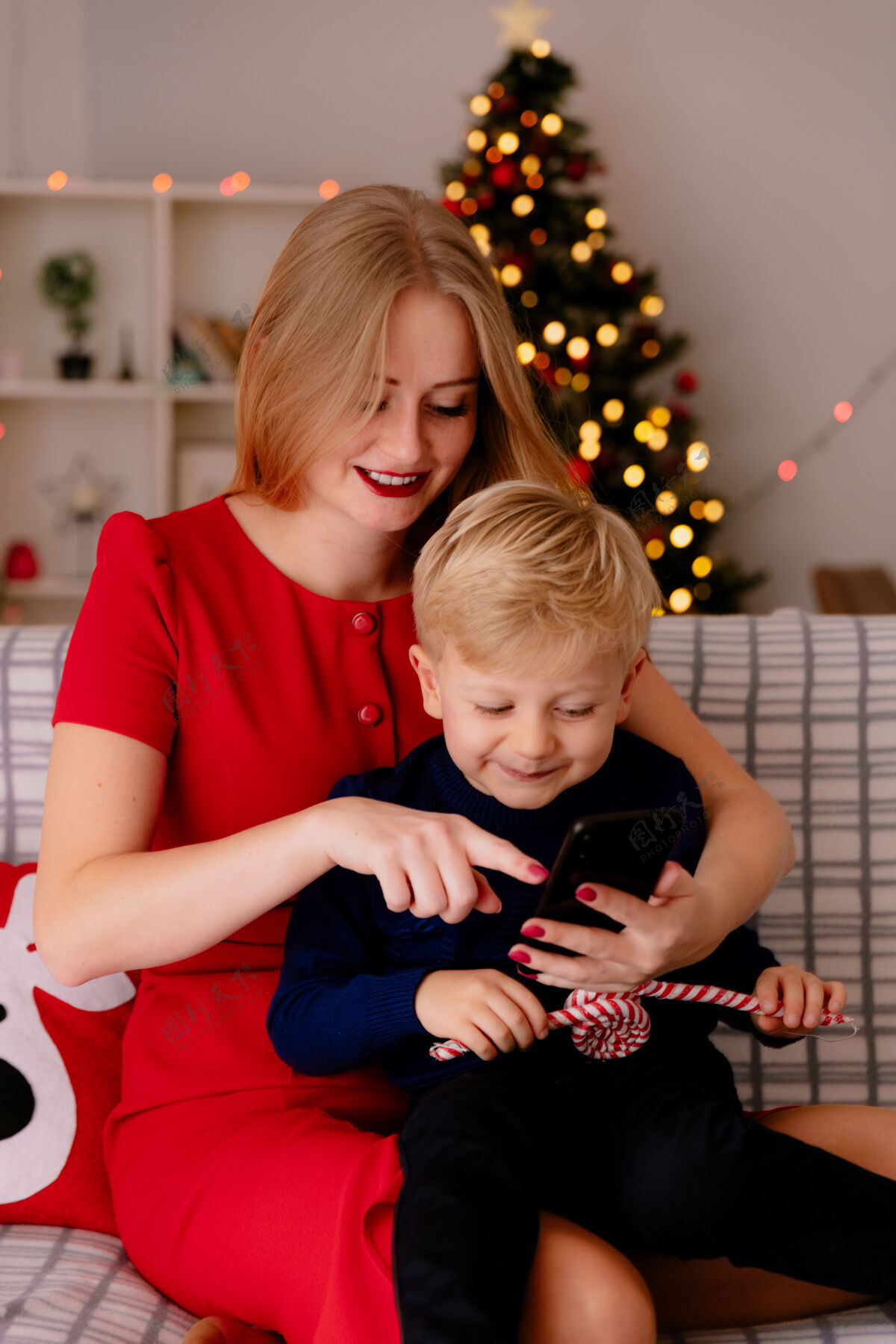 母亲在一间以圣诞树为背景的装饰房间里 穿着红色连衣裙的快乐妈妈和她的孩子坐在沙发上 智能手机放在沙发上连衣裙快乐沙发