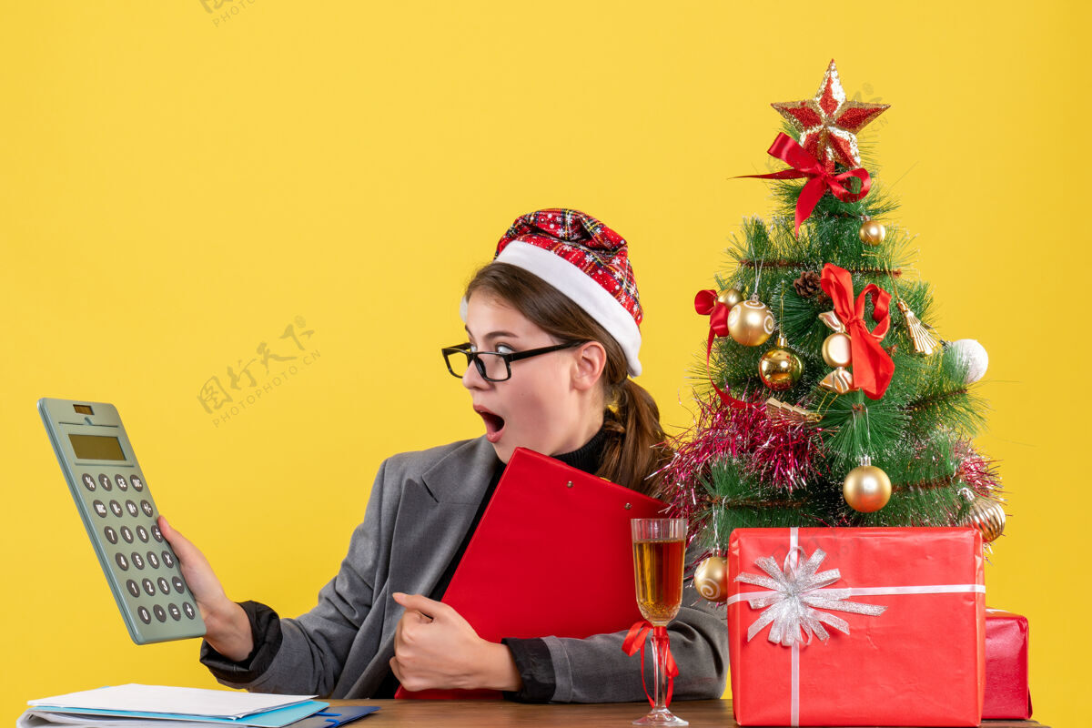 计算器前视图震惊的女孩与圣诞帽坐在桌边看计算器圣诞树和礼物鸡尾酒看女孩人