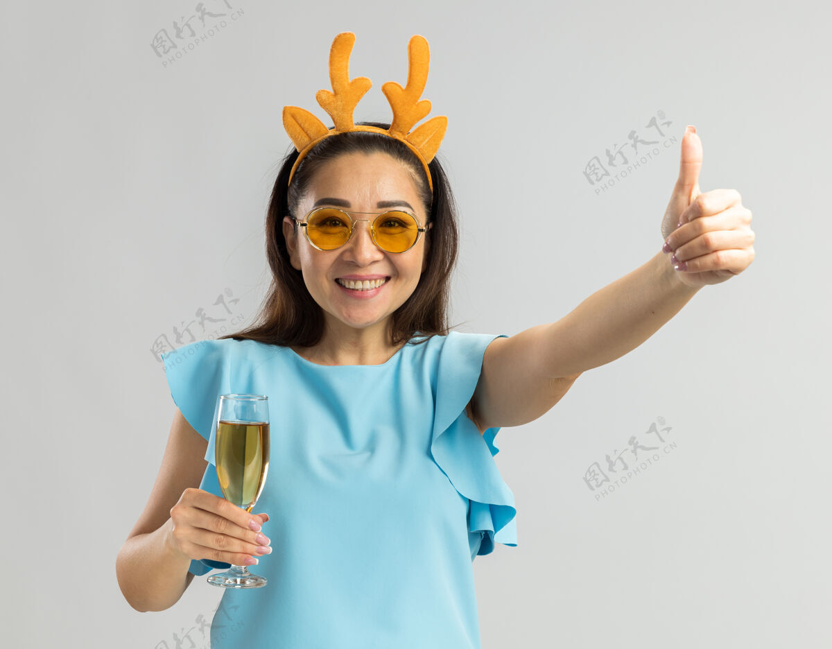 女人身穿蓝色上衣的年轻女子 戴着滑稽的鹿角边 戴着黄色的玻璃杯 手里拿着一杯香槟 脸上露出愉快的笑容 竖起大拇指鹿举着圣诞