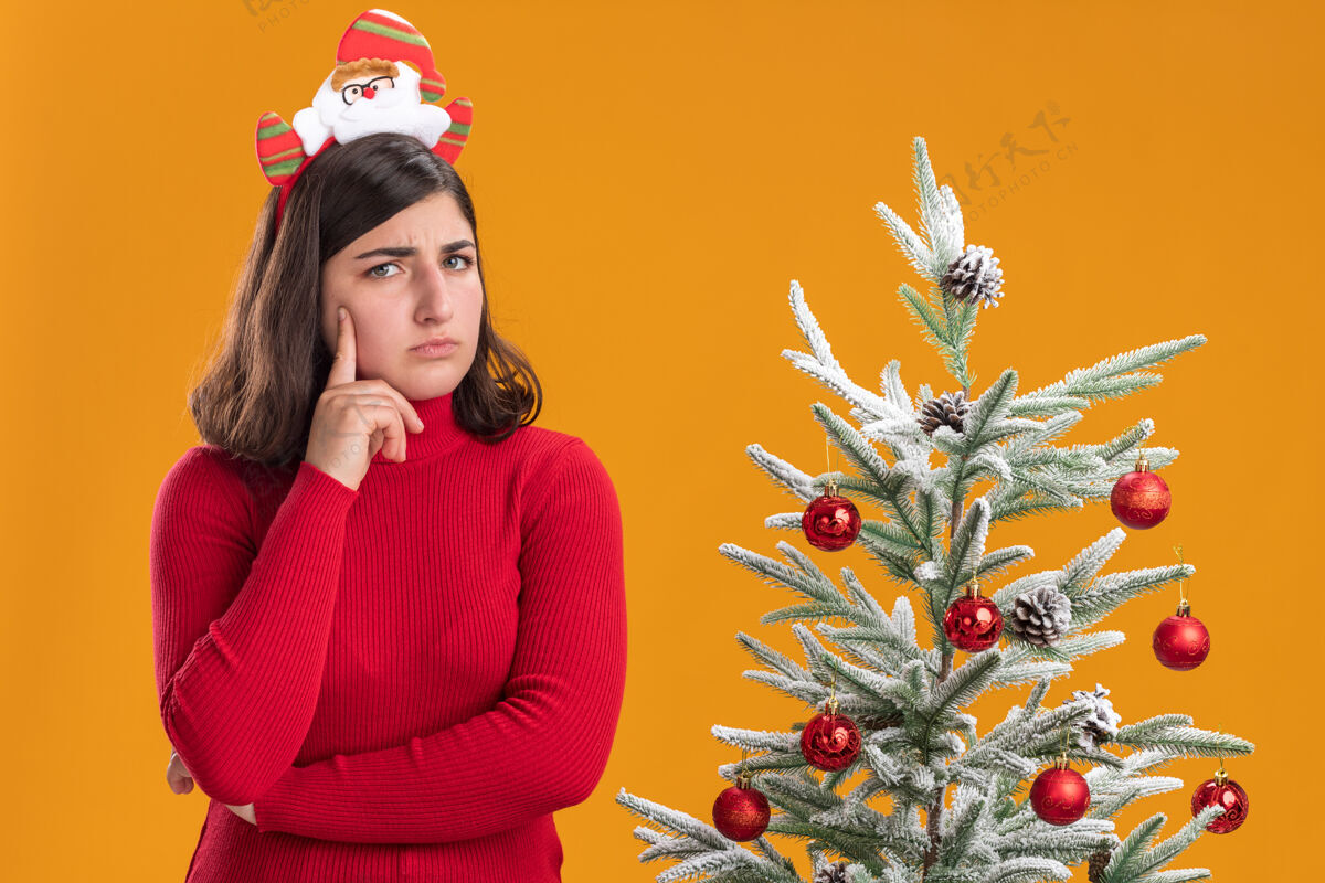 相机穿着圣诞毛衣的年轻女孩戴着滑稽的头带 看着橙色背景下圣诞树旁的摄像机头带站年轻