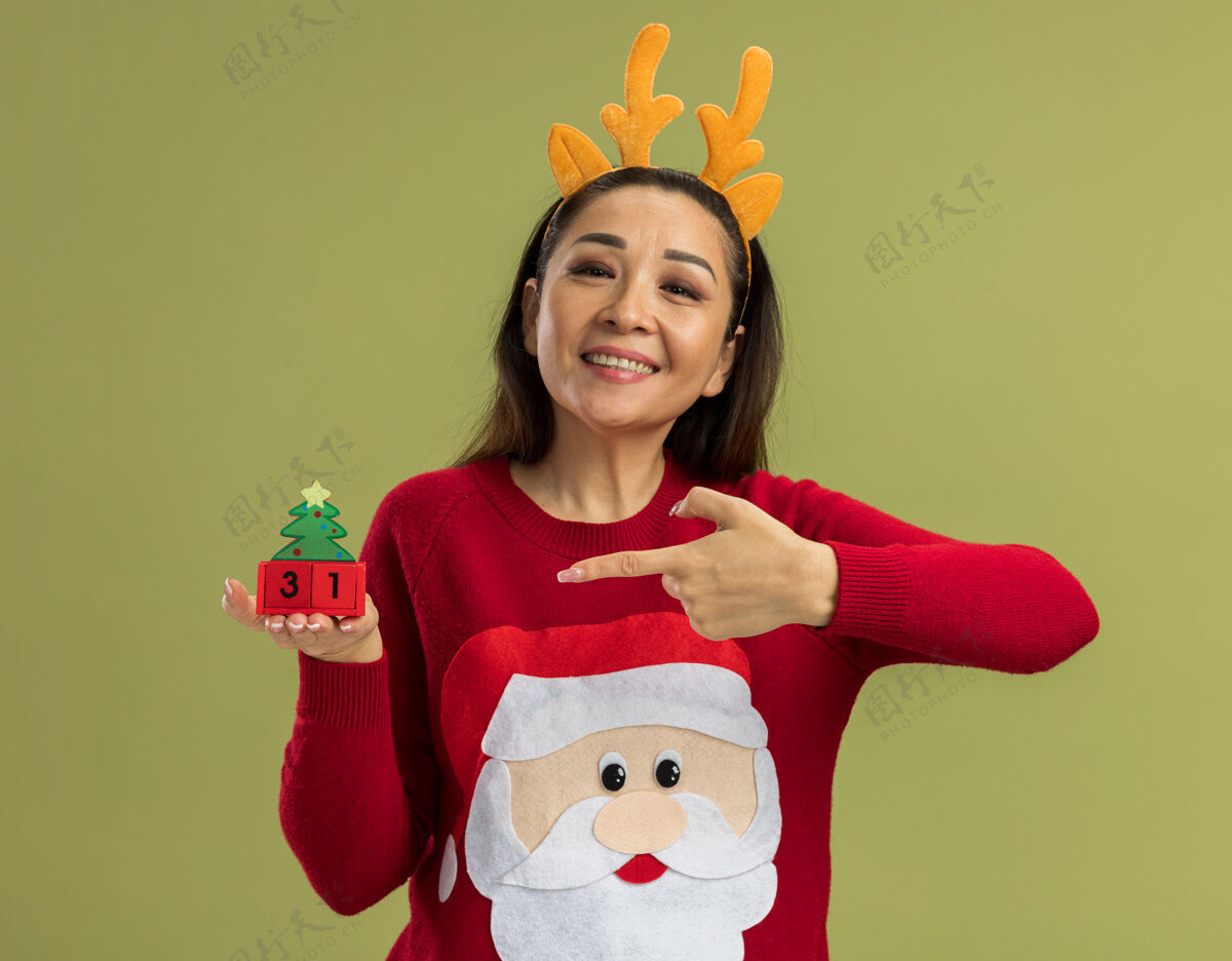 立方体穿着红色圣诞毛衣的快乐年轻女子 戴着有趣的鹿角边 展示着新年日期的玩具立方体 她愉快地笑着 用食指指着站在绿色背景上的立方体站立日期毛衣