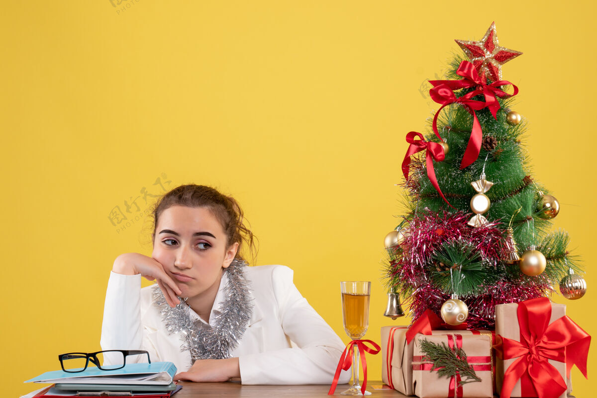 圣诞节前视图女医生围坐在圣诞礼物和圣诞树周围 在黄色背景下感到无聊无聊情绪办公室