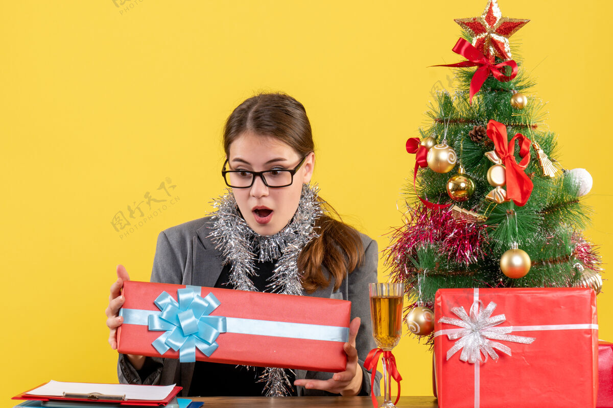 坐着正面图戴着眼镜的大眼睛女孩坐在桌子旁看着她的圣诞树和鸡尾酒礼物圣诞节桌子微笑