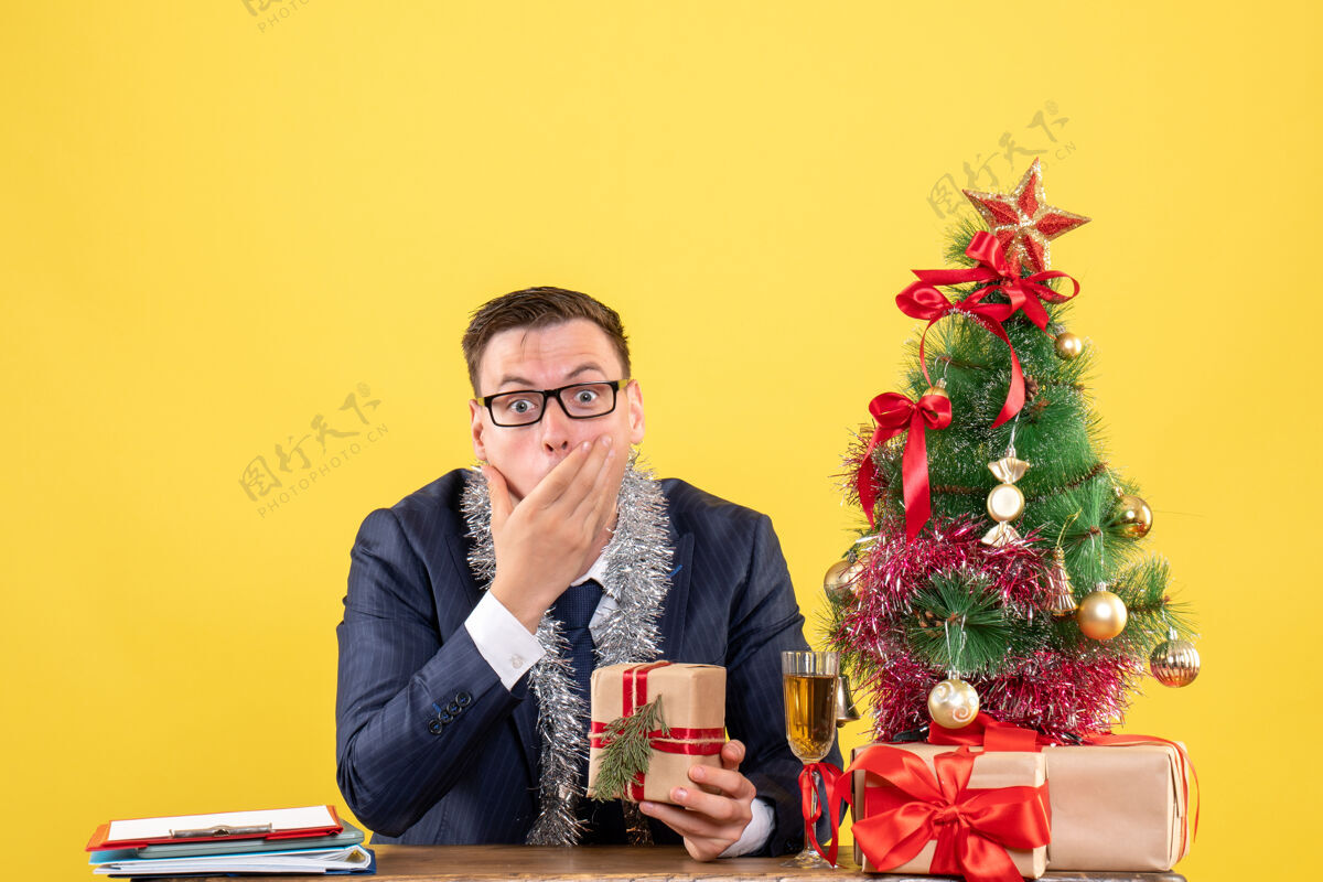 男性一个好奇的男人把手放在嘴上 坐在圣诞树旁的桌子旁 用黄色的礼物呈现成人礼物嘴巴