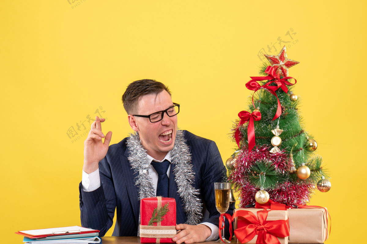 运气正面图：快乐的商人在圣诞树旁的桌子旁做着好运的手势 黄色的礼物树新郎男人