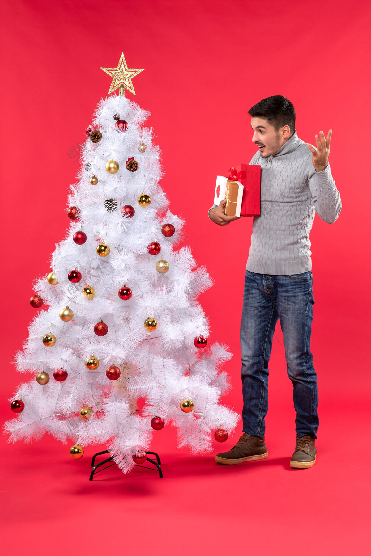 圣诞英俊的年轻人站在装饰过的白色新年树旁 手里拿着他的礼物礼物礼物圣诞树