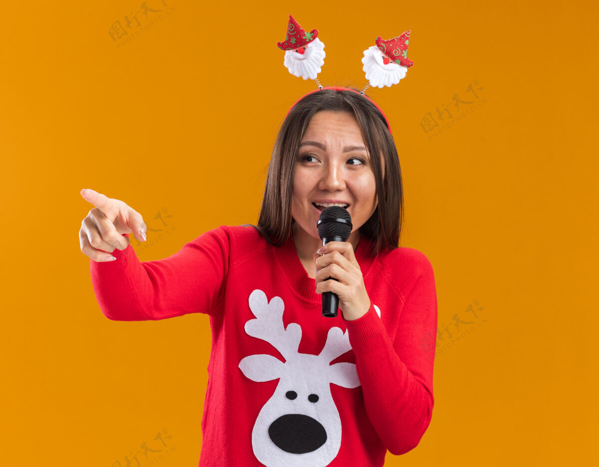 女人笑容满面的年轻亚洲女孩戴着圣诞发箍 穿着毛衣 对着麦克风讲话 指着隔离在橙色墙上的一侧麦克风箍边