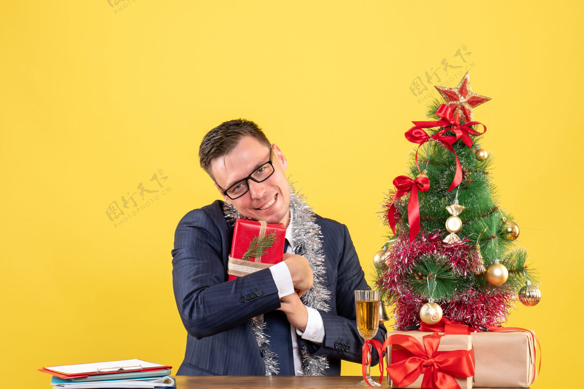 肖像正面图：幸福的男人紧紧地拿着礼物坐在圣诞树旁的桌子旁 黄色的礼物摆在桌子上坐着时尚人