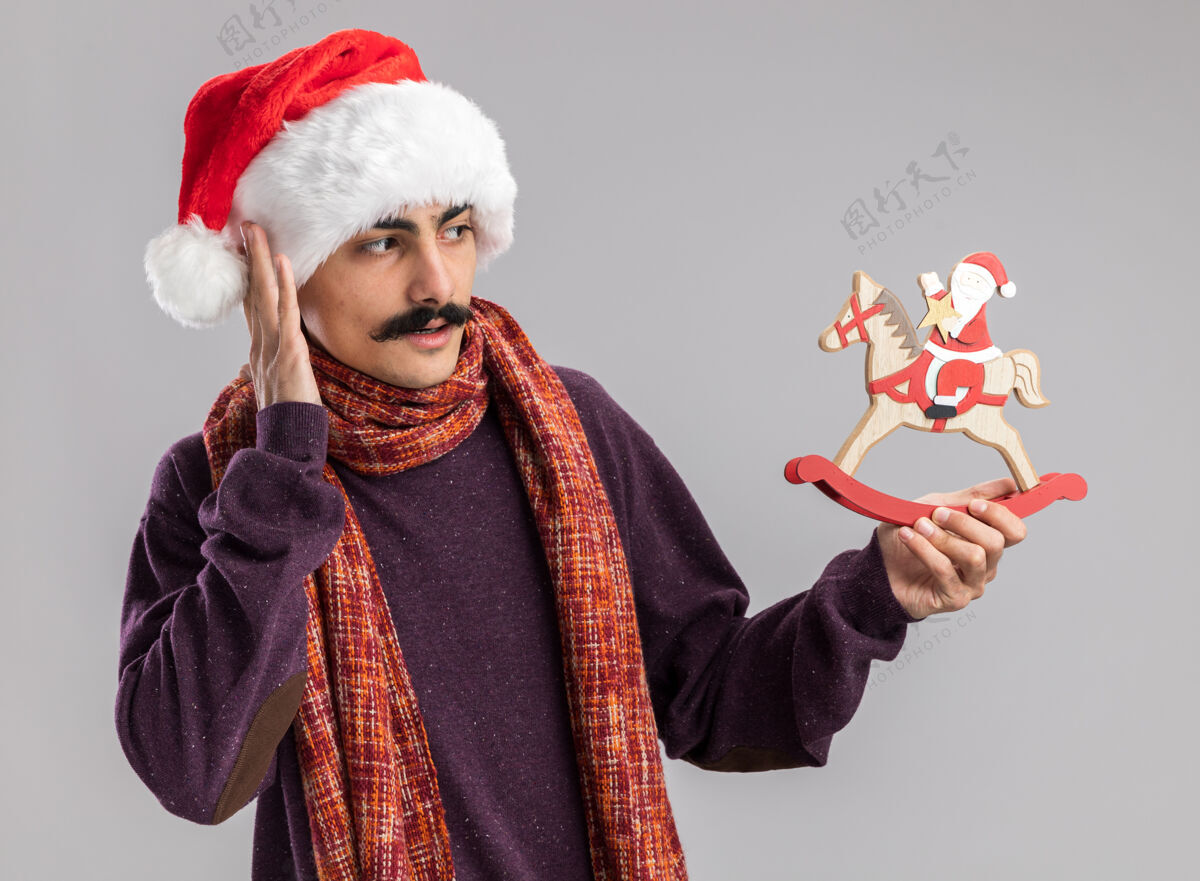 圣诞快乐小胡子男人戴着圣诞老人的帽子 脖子上围着暖和的围巾 手里拿着圣诞玩具 困惑地看着它快乐圣诞温暖