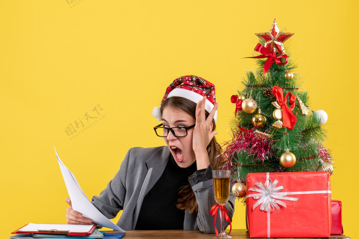 礼物前视图震惊女孩与圣诞帽坐在桌边看文件圣诞树和礼物鸡尾酒女孩圣诞节纸