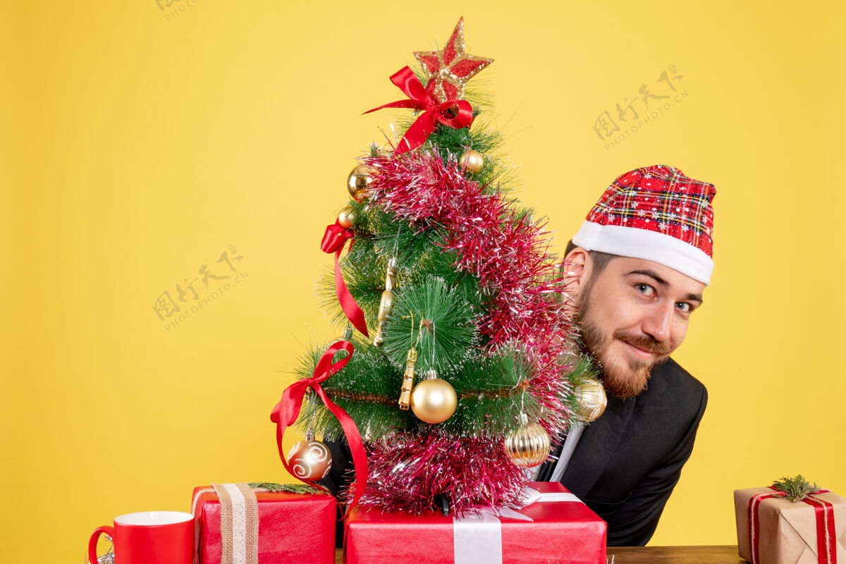 圣诞节正面图：坐在黄色办公桌上拿着礼物的男员工圣诞办公室彩色新年礼物庆祝男树
