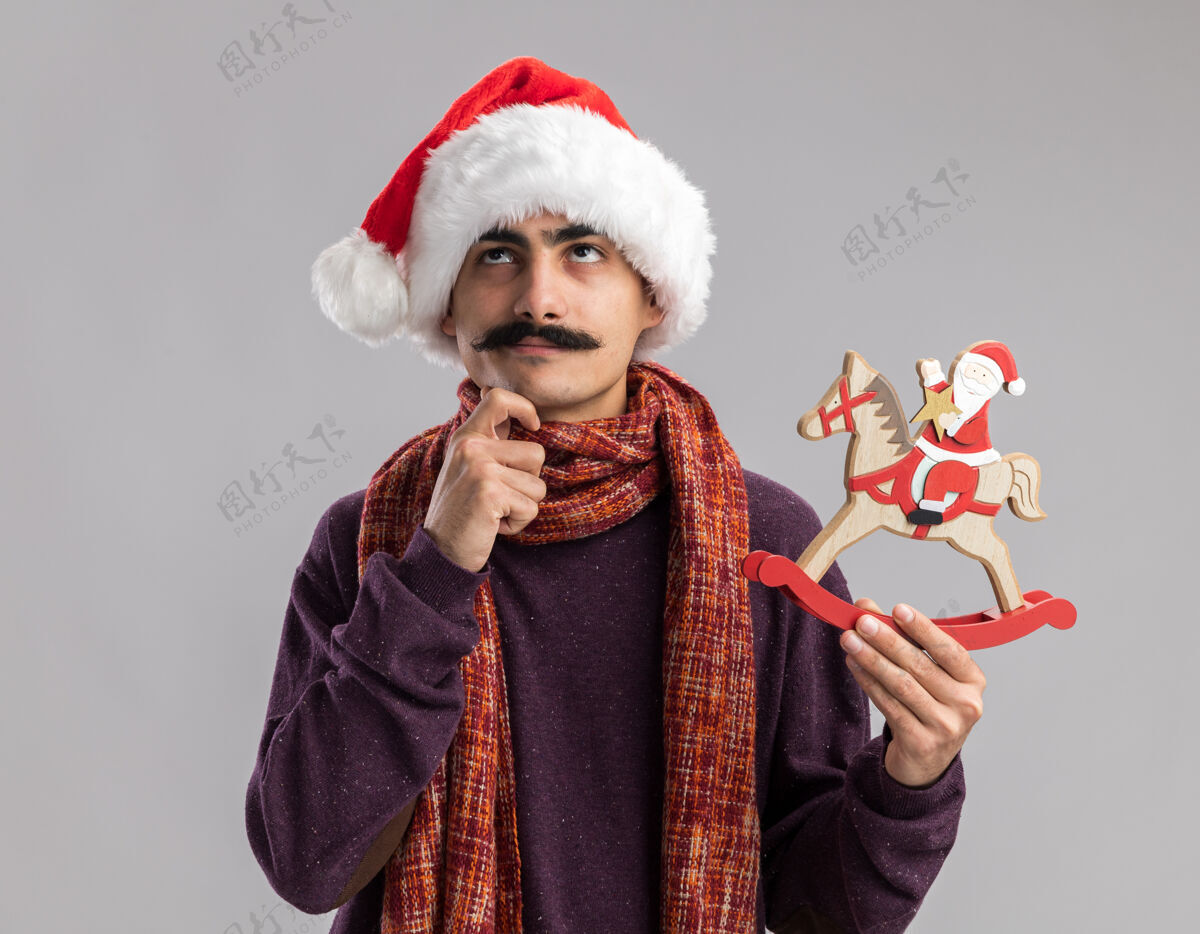 温暖年轻的胡子男人戴着圣诞老人帽 脖子上围着暖和的围巾 手里拿着圣诞玩具 站在白色的背景上困惑地看着困惑圣诞帽子