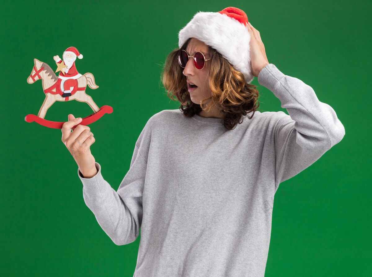 帽子年轻人戴着圣诞老人帽 戴着红眼镜 手里拿着圣诞玩具 困惑地看着它 手放在头上 站在绿色的背景上站着头眼镜