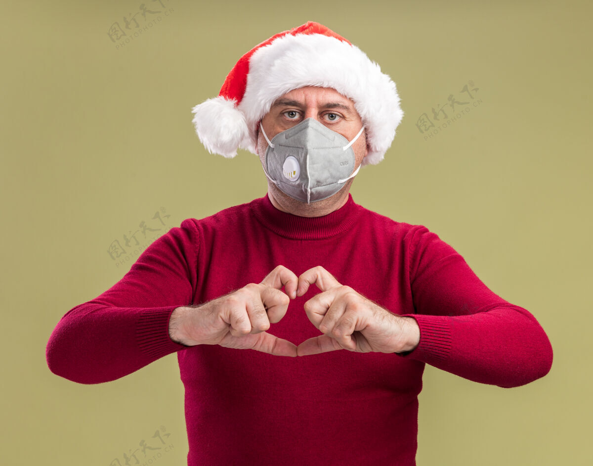 手势中年男子戴着圣诞老人帽戴着护面面具看着镜头严肃的做脸用手指站在绿色的背景上做着心形手势年龄圣诞老人面部