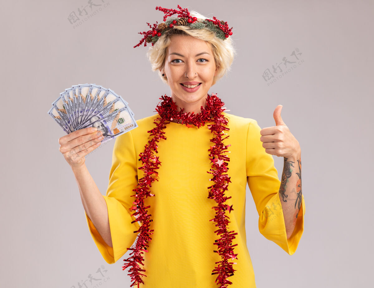圣诞节微笑着的金发女郎戴着圣诞花环 脖子上戴着金属丝花环 手里拿着钱 看着相机 在白色背景上孤立地竖起大拇指钱拇指举行