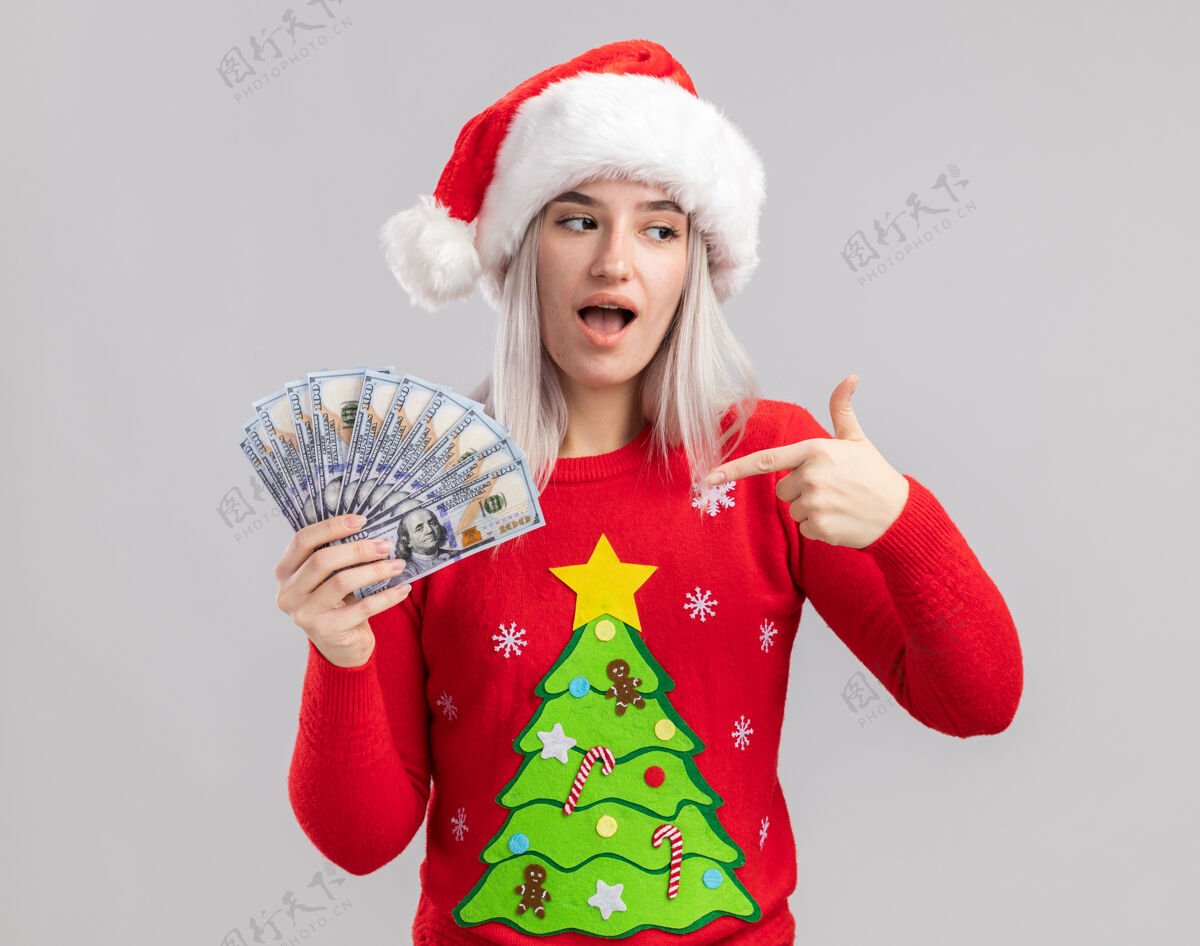 圣诞快乐穿着圣诞毛衣 戴圣诞帽的金发女郎拿着现金 用食指指着钱 又高兴又惊讶手指女人现金