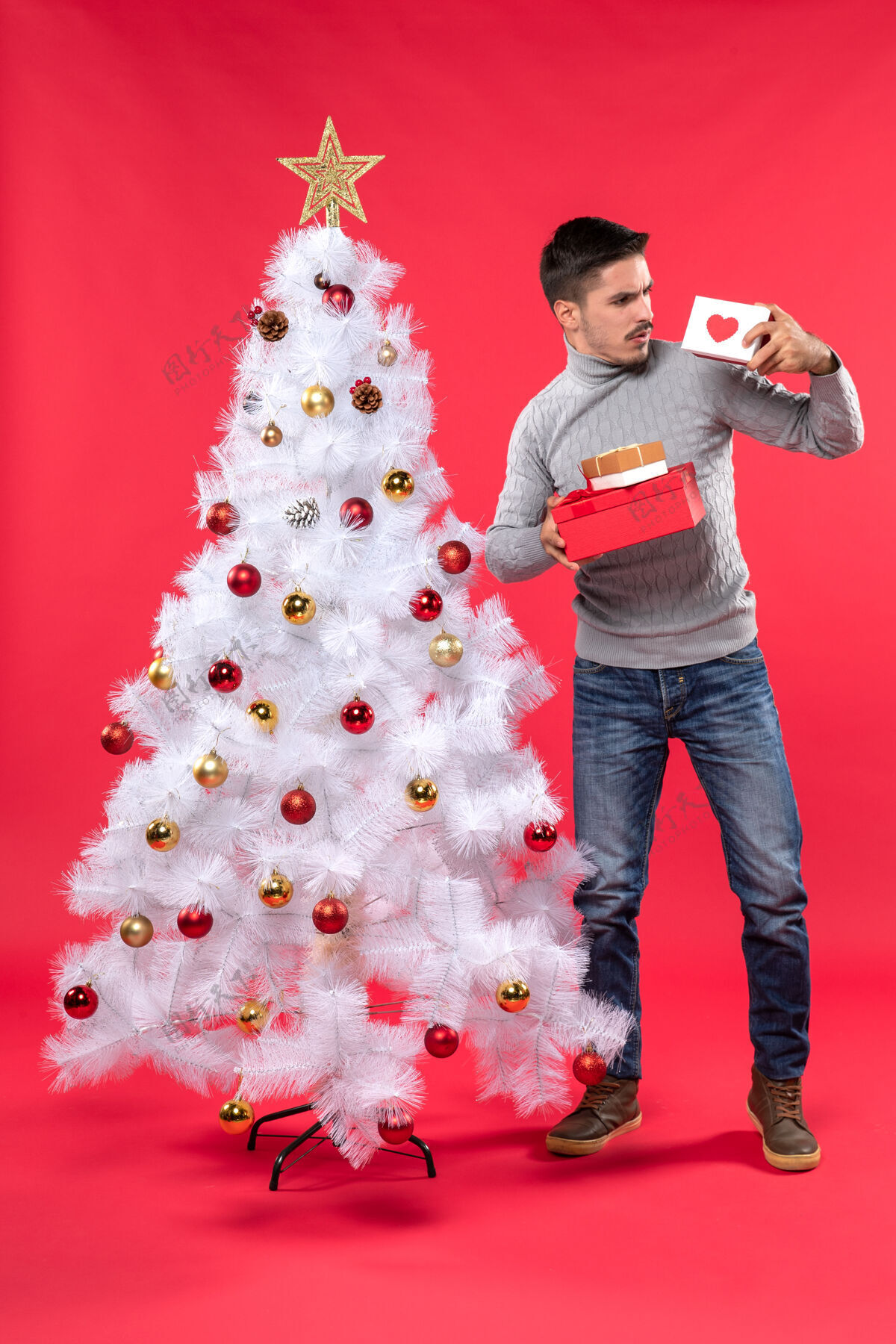 漂亮困惑的年轻人穿着灰色上衣站在装饰过的圣诞树旁 手里拿着红色的礼物年轻人可爱礼物