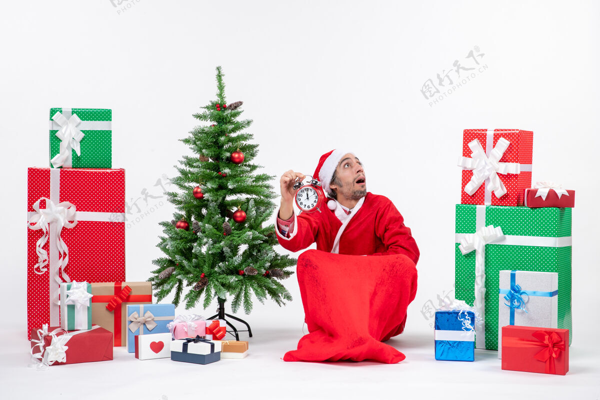 快乐圣诞老人带着惊讶的表情坐在礼品盒和圣诞树旁展示表情地