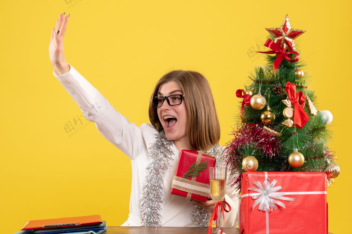 抱着正面图：女医生坐在桌子前 手里拿着黄色背景的礼物 还有圣诞树和礼品盒人物年医生