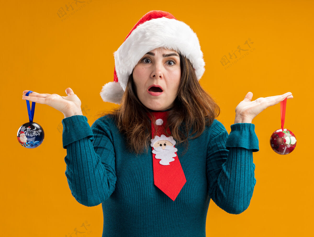 震惊震惊的成年白人妇女与圣诞帽和圣诞领带举行玻璃球饰品孤立在橙色背景与复制空间橙色帽子领带