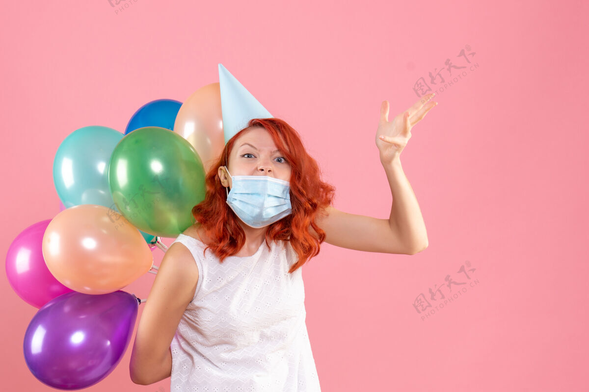 五颜六色正面图是粉红色墙上戴着消毒面具手持彩色气球的年轻女子快乐快乐人们